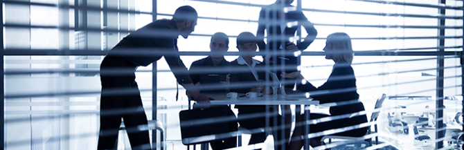 Looking through venetian blinds into boardroom, five people are having a meeting